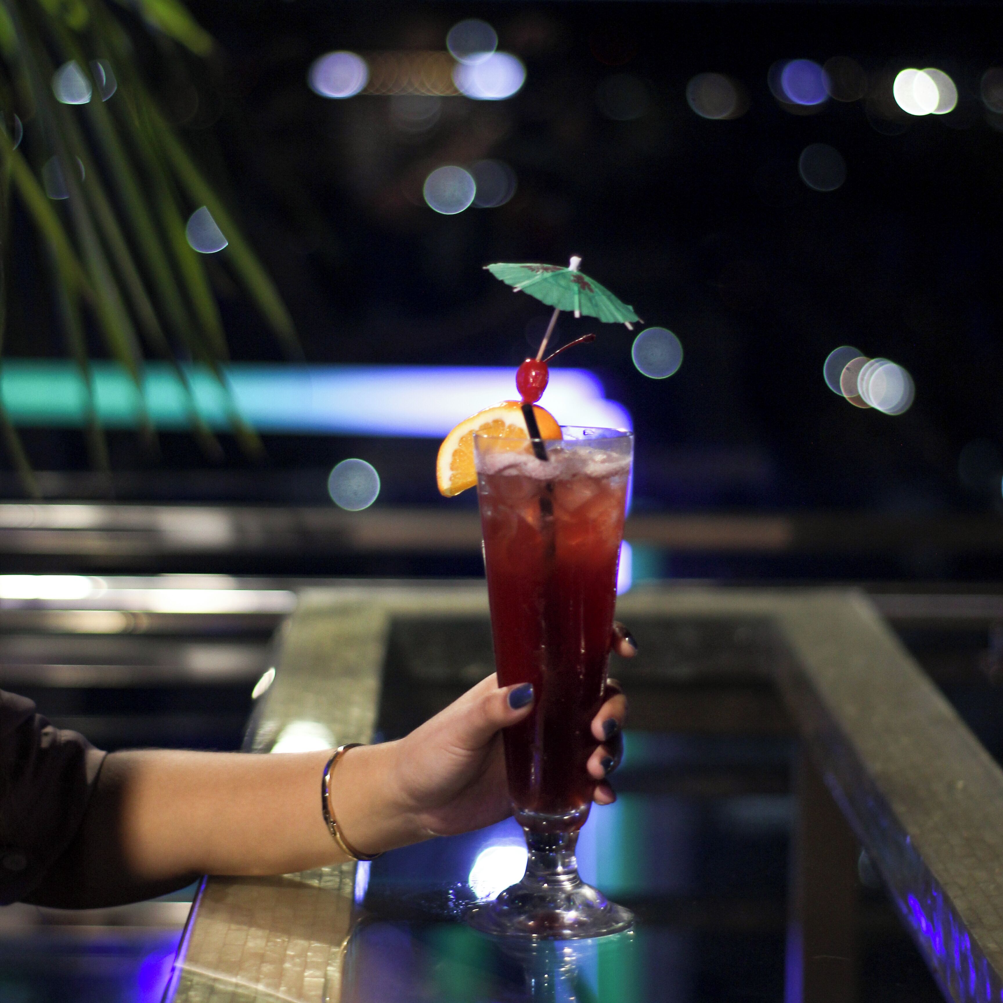 A hand holding a tall glass with a red beverage, garnished with an orange slice and a cherry, and an umbrella.