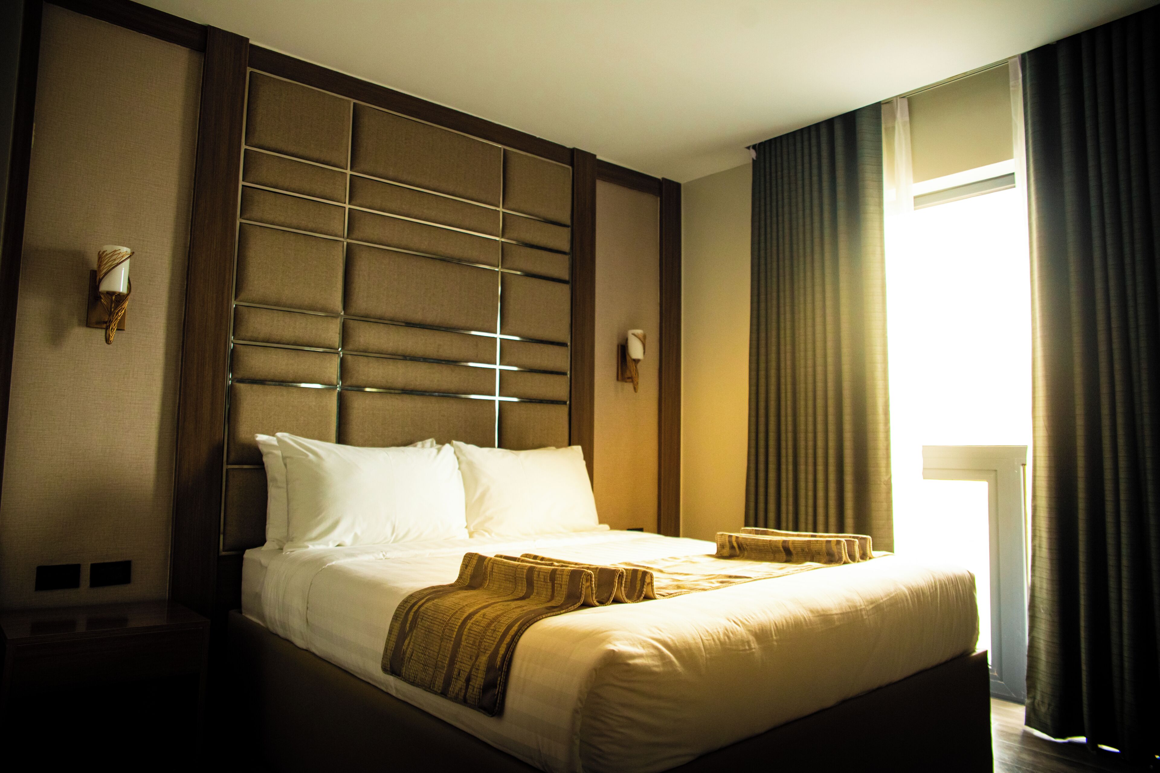 A hotel room with a large bed, a headboard with a grid pattern, bedside lamps, and a window with curtains.