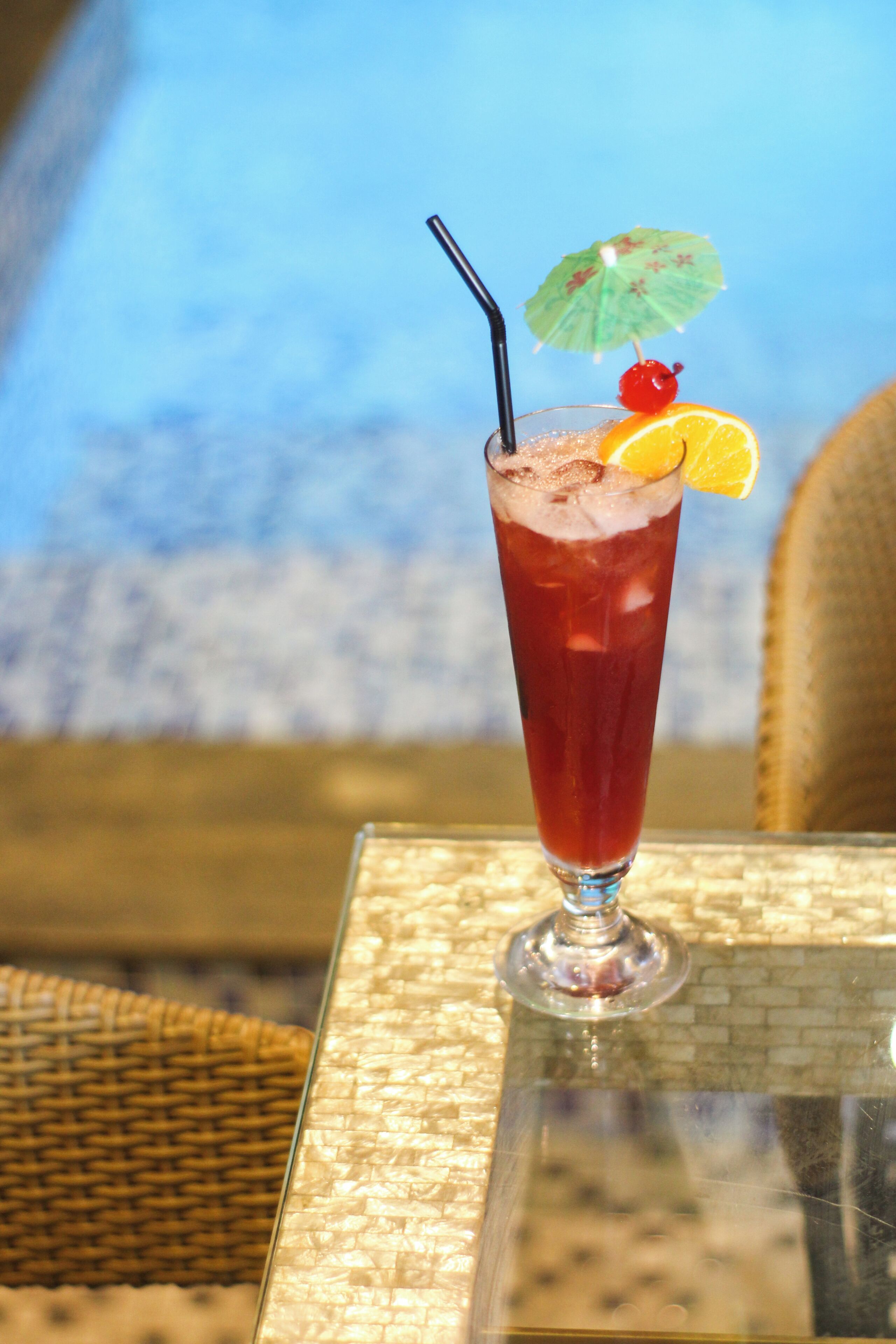 A tall glass with a red beverage, a straw, a cherry, and an orange slice on a table with a woven chair and a view of the sky.