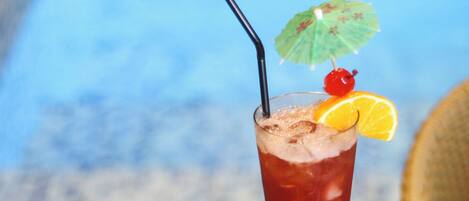 A tall glass with a red beverage, a straw, a cherry, and an orange slice on a table with a woven chair and a view of the sky.