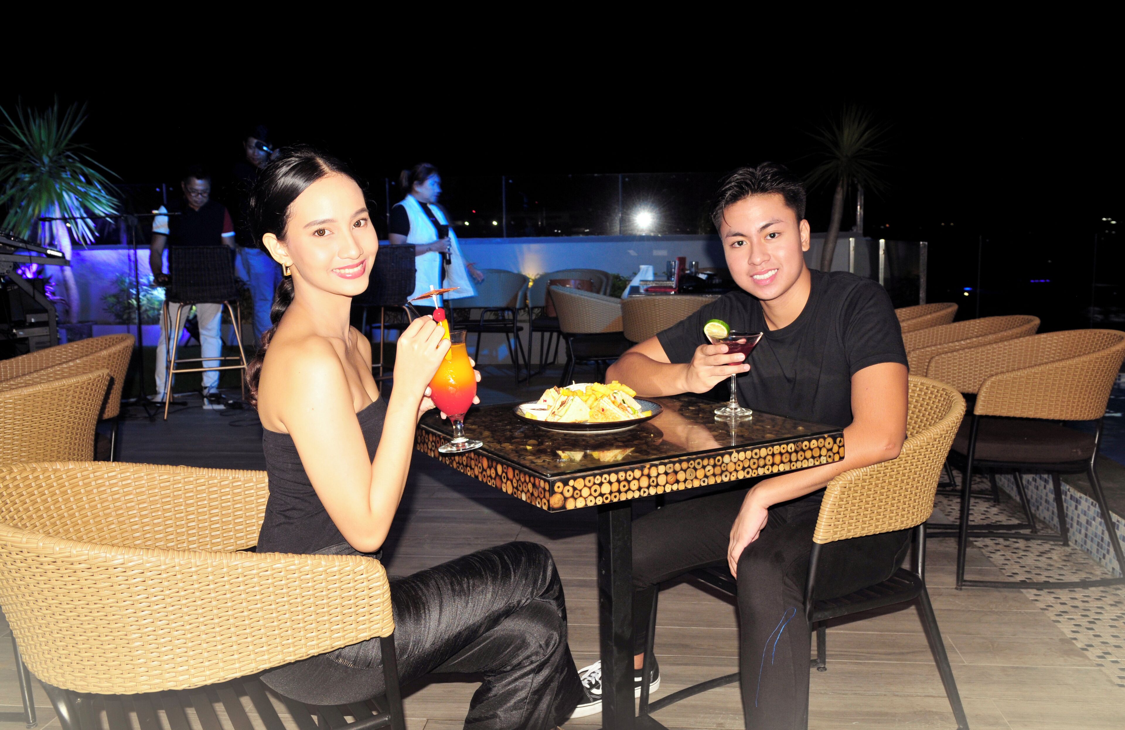 Two people seated at a table with wicker chairs, a plate of food, and a drink.