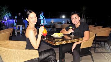 Two people seated at a table with wicker chairs, a plate of food, and a drink.
