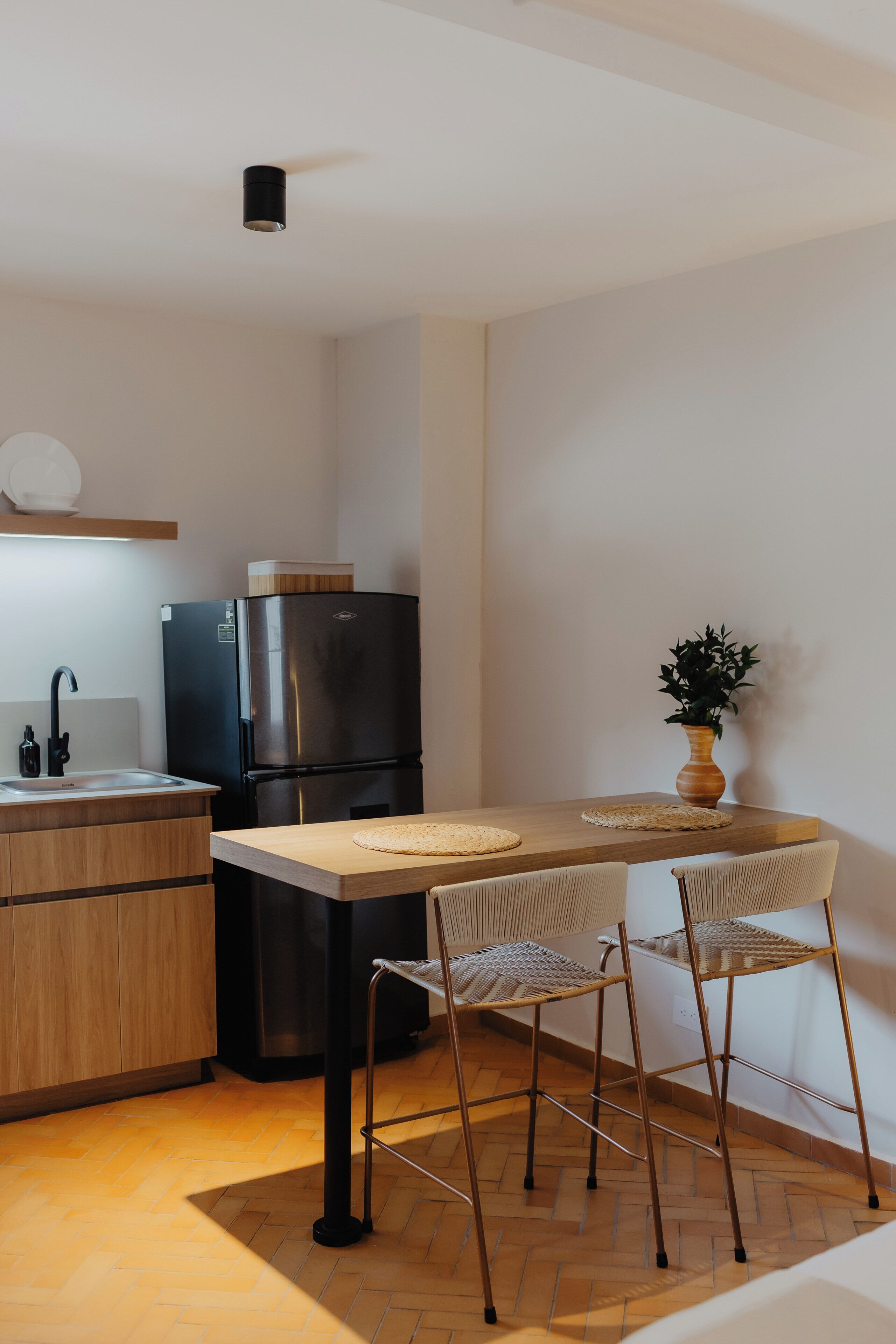 Een kleine keuken met een zwarte koelkast, houten kastjes, een wastafel en twee rieten stoelen.