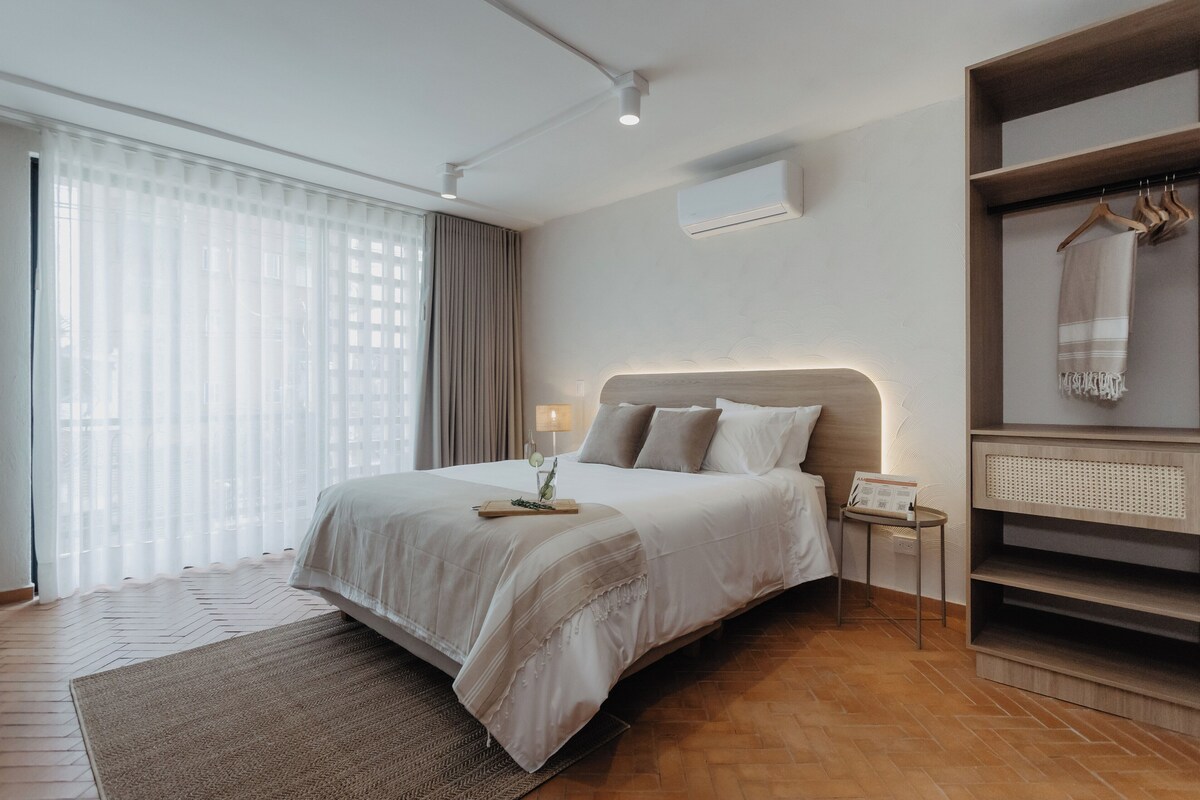 A modern hotel room with a large bed, a bedside table with a lamp, a small side table, and a built-in wardrobe with a mirror.