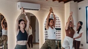 Una clase de yoga en una sala amplia con vigas de madera, ventanas grandes y una lámpara colgante.