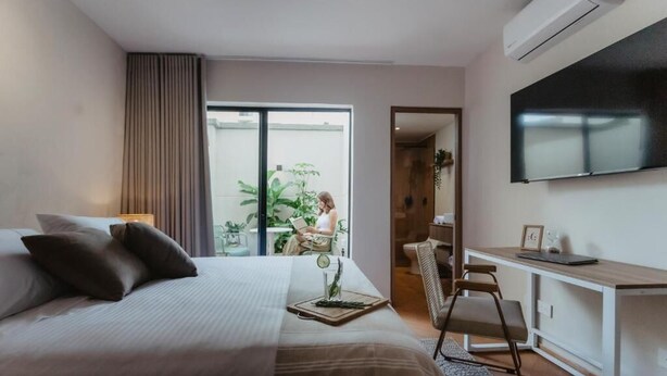 A modern hotel room with a bed, desk, TV, and a view of a balcony with a person reading. - Pranik Living Co-mmunity (Medellín)