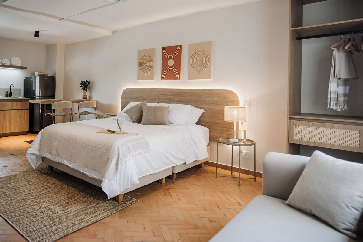 A modern hotel room with a large bed, a bedside table, a chair, a sofa, a small kitchen area, and a wardrobe.