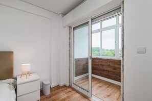 Ein Schlafzimmer mit Bett, Nachttisch, Lampe und einer Schiebetür zu einem Balkon mit Blick ins Grüne.