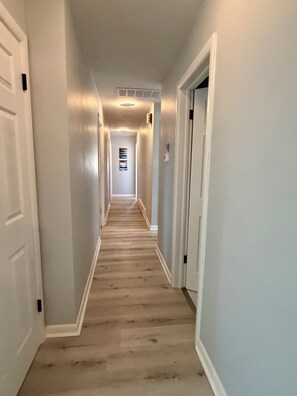 Un couloir aux murs blancs et au plancher en bois, qui mène à une porte au fond.
