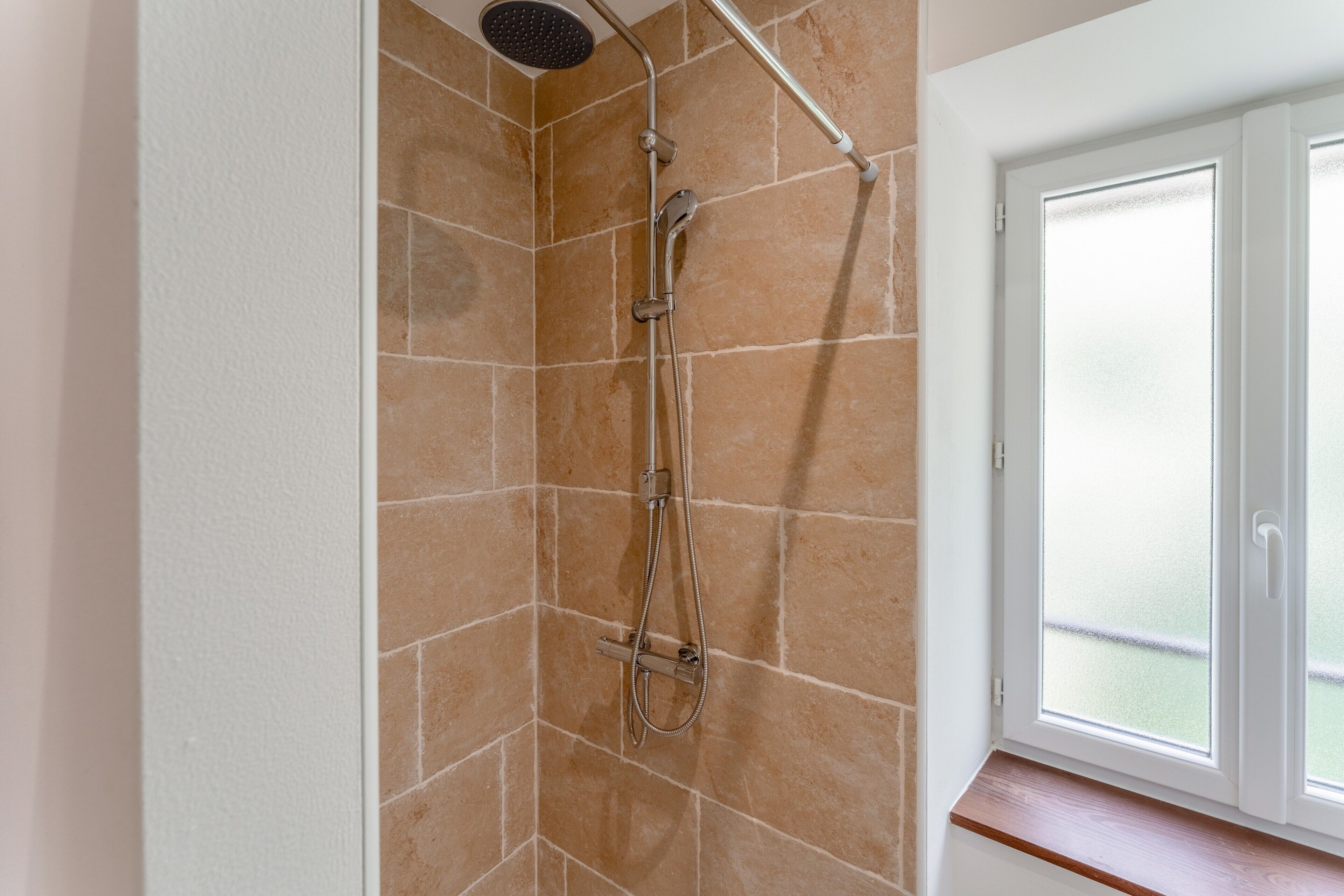 Een douche met een handdouche en een extra douchestraal, een raam met uitzicht op een balkon, en een witte deur.