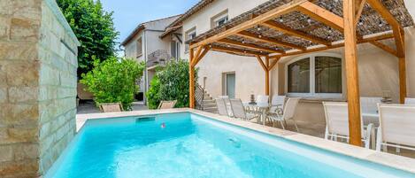 Una piscina amb una pèrgola de fusta, mobiliari exterior i una paret de pedra.