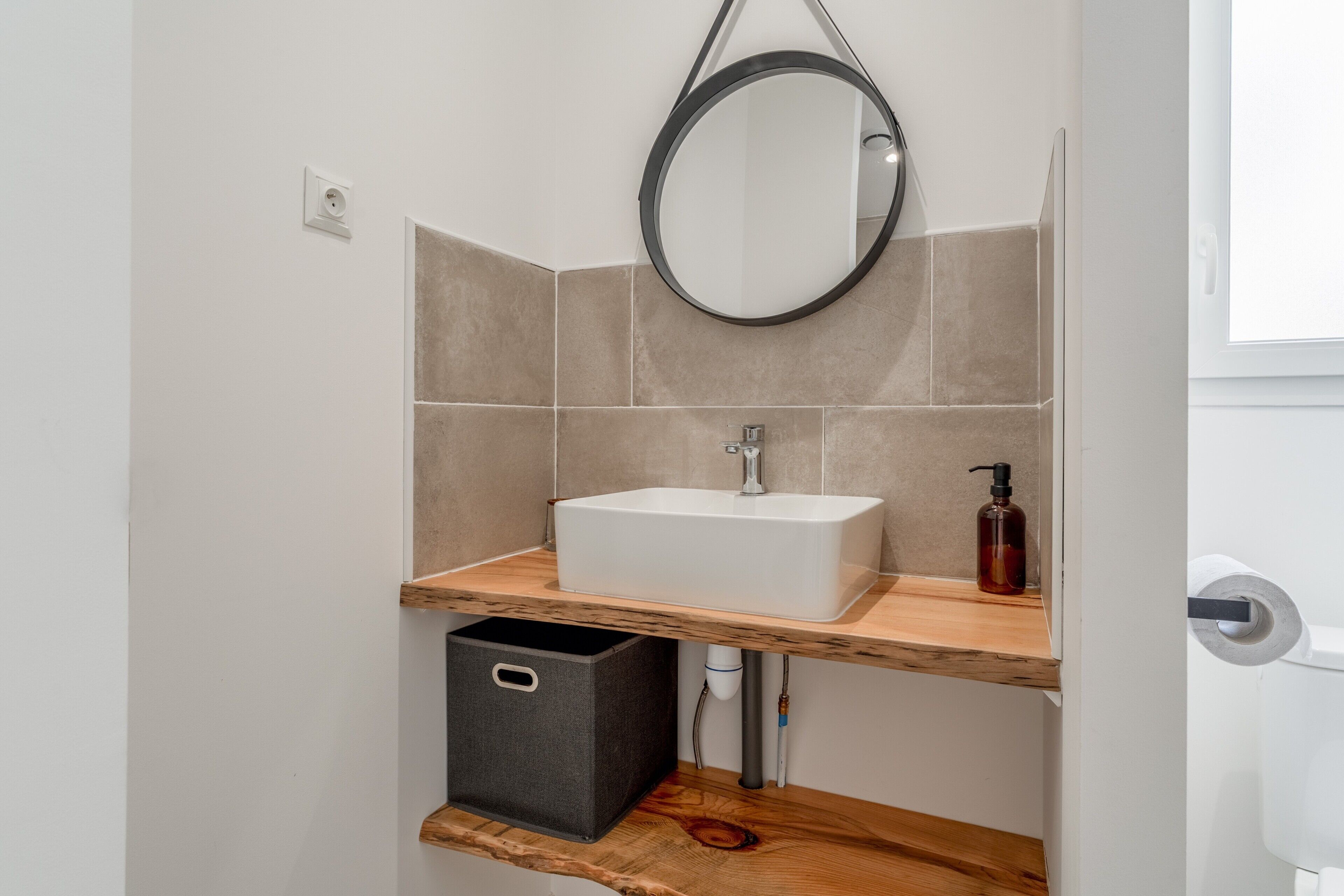 A bathroom with a white sink, a mirror, beige tiles, a wooden shelf, and a black storage box.