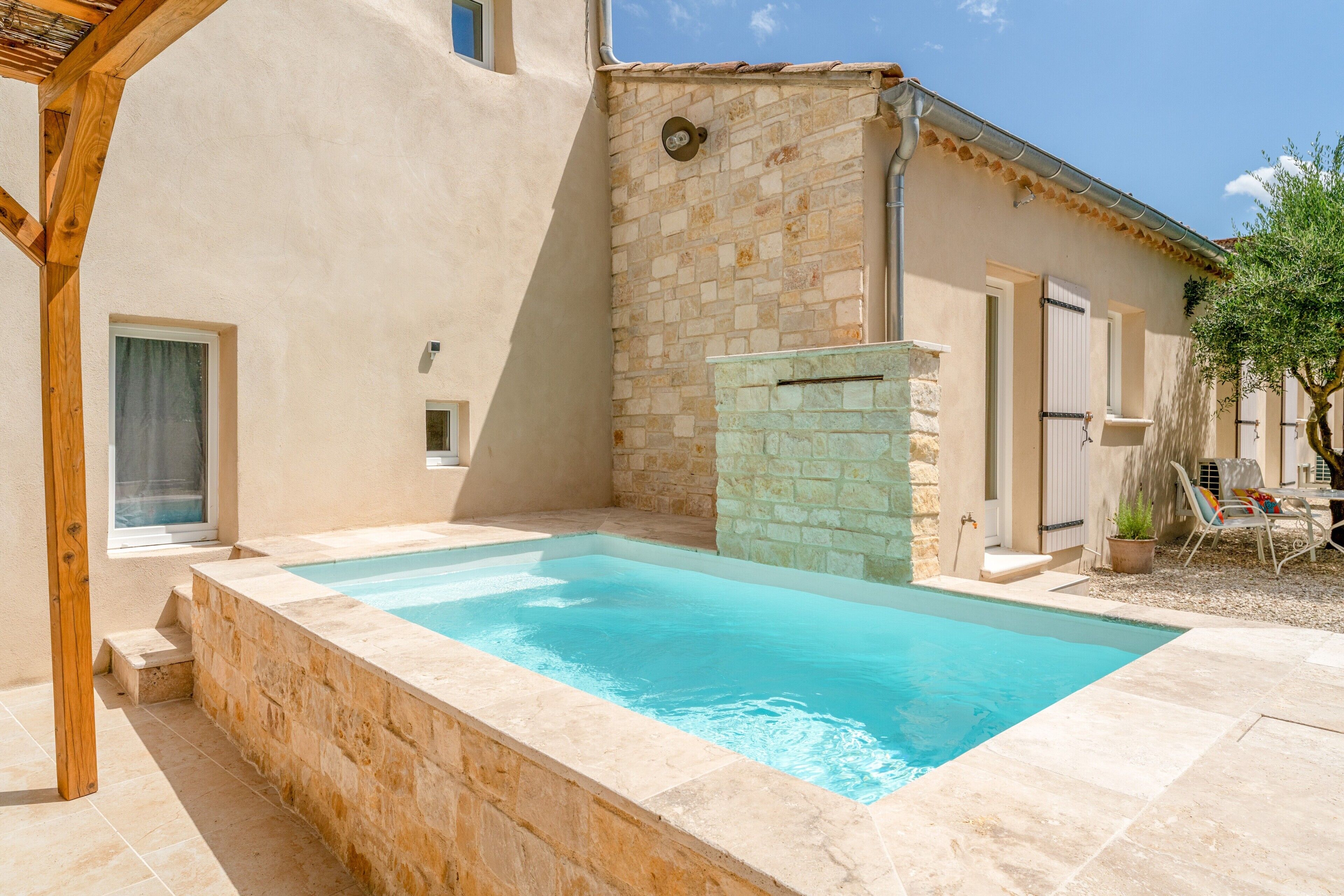 A swimming pool with a stone wall, a wooden pergola, and a building with a tiled roof.