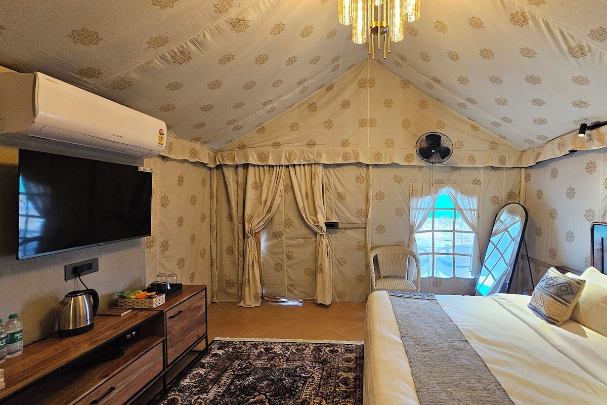 Habitación tipo tienda de campaña con una cama, televisión, ventilador y ventana con cortinas.