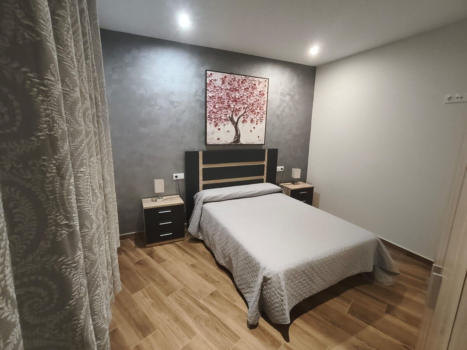 A bedroom with a bed, nightstands, a wall-mounted painting, and a curtain.