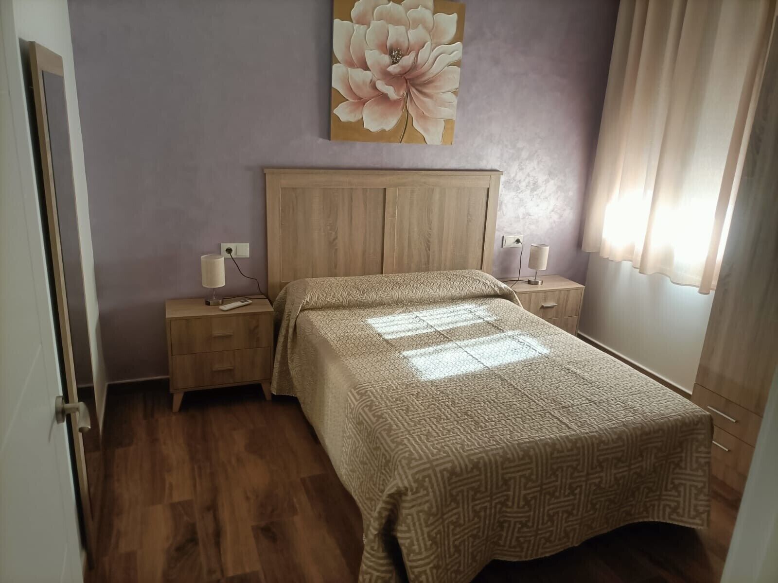 Ein Schlafzimmer mit einem Holz-Kopfteil, einer gemusterten Bettdecke, Nachttischlampen und einem an der Wand hängenden Blumenbild.