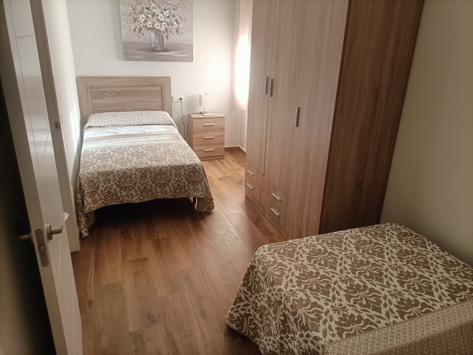 A bedroom with two beds, a wardrobe, a bedside table, and a wall-mounted lamp.