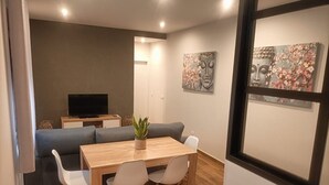 A modern living room with a sofa, a dining table, and chairs. There is a television on a stand and artwork on the wall.