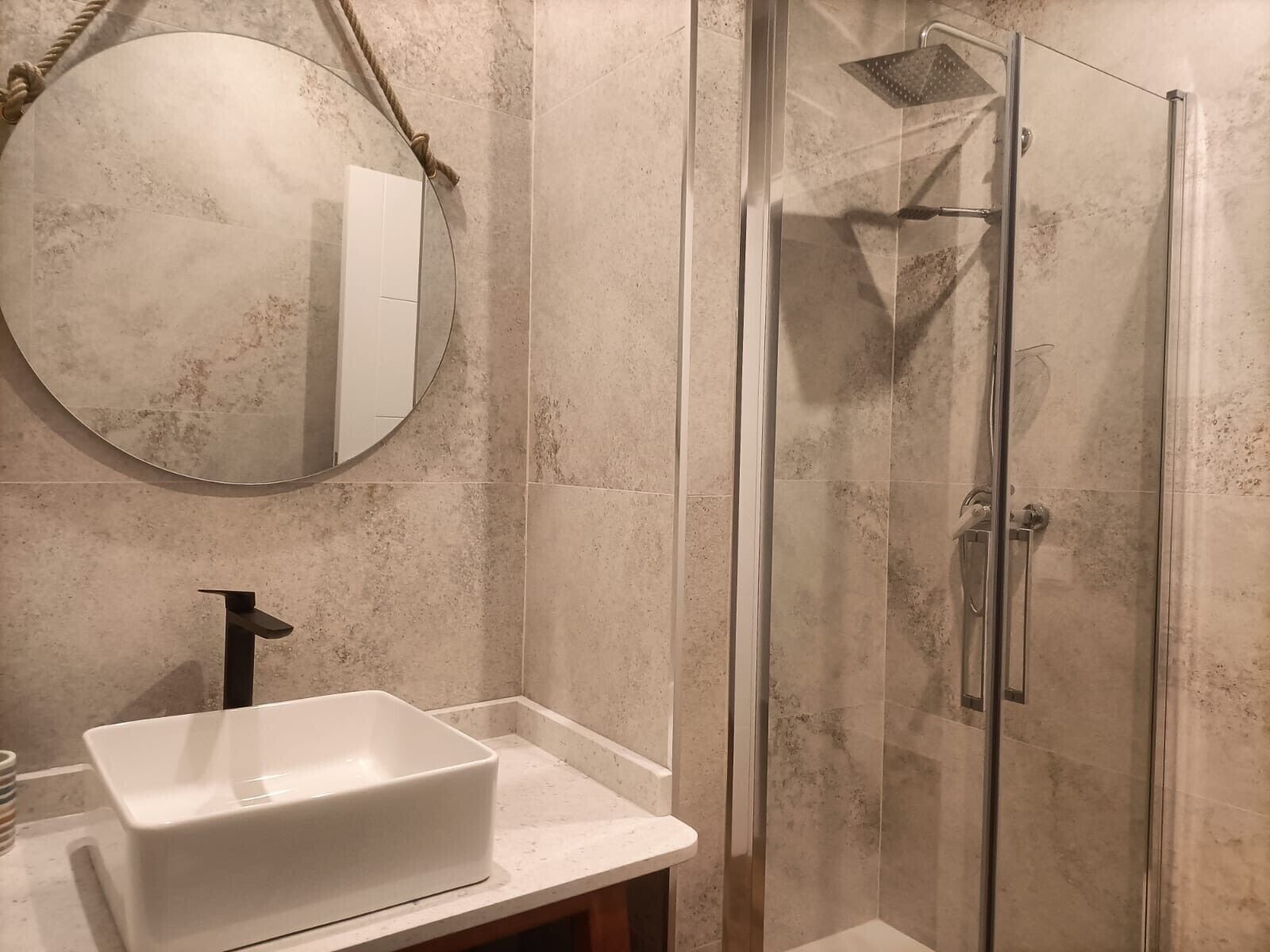 A bathroom with a glass-enclosed shower, a white sink, and a round mirror with a rope.