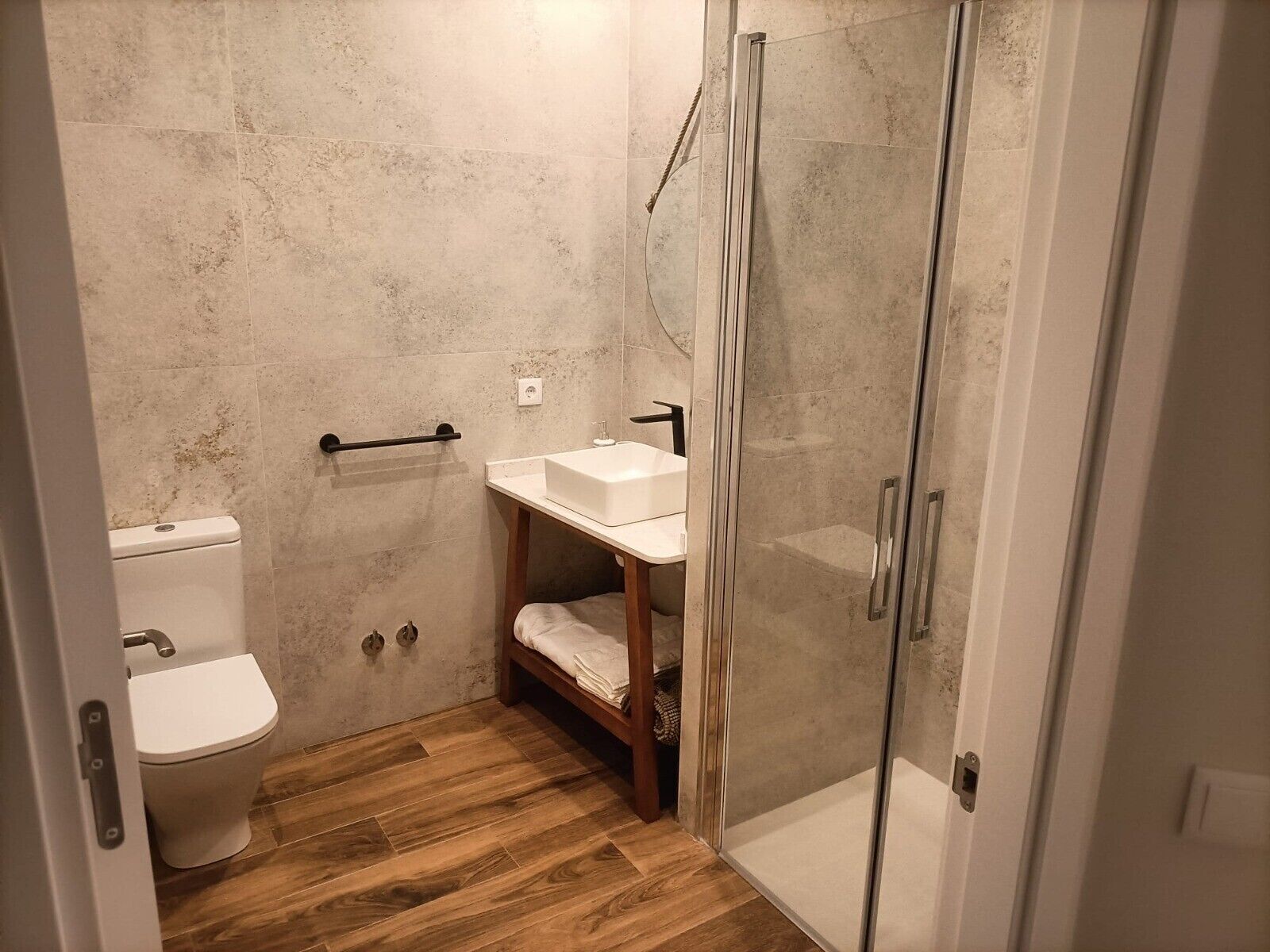 Ein Badezimmer mit Toilette, Waschbecken, Dusche mit Glastür und Holzregal.