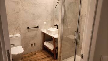 Ein Badezimmer mit Toilette, Waschbecken, Dusche mit Glastür und Holzregal.