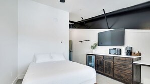 A bedroom with a bed, a TV, a small kitchenette, and a door. - The Lawson District | Marigold Suite (Midlothian)