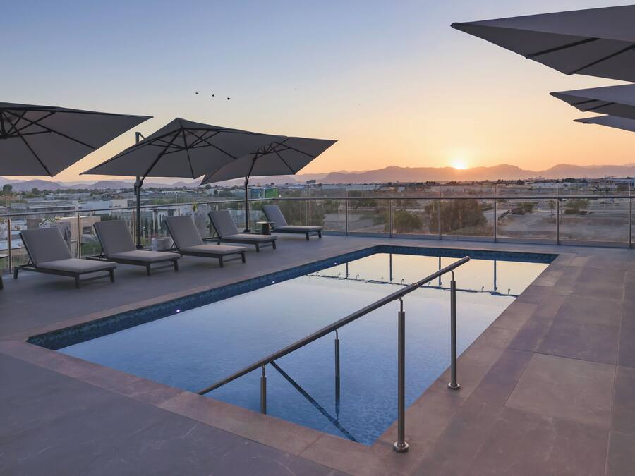 A rooftop pool area with lounge chairs and umbrellas, overlooking a cityscape and mountains.