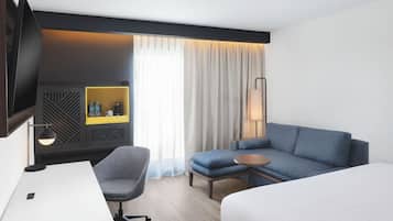 Habitación de hotel con cama, escritorio, silla, sofá, televisor, lámpara, mesita de noche y ventana con cortinas.