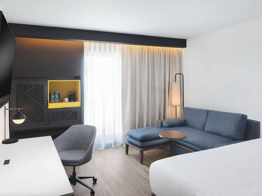 Habitación de hotel con cama, escritorio, silla, sofá, televisor, lámpara, mesita de noche y ventana con cortinas.