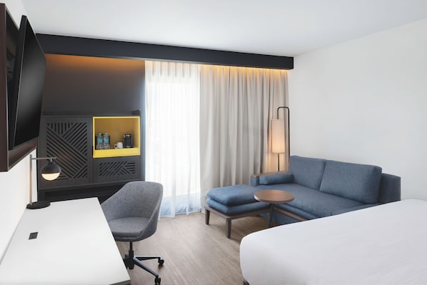 Une chambre d’hôtel comprenant un lit, un bureau, une chaise, un canapé, une télévision, une lampe, une table de chevet et une fenêtre avec des rideaux.