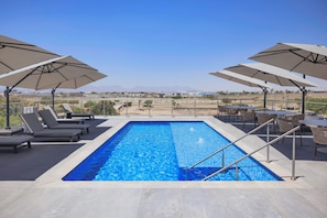 Une piscine rectangulaire à l’eau d’un bleu limpide, entourée de chaises longues et de parasols, et offrant une vue sur des montagnes lointaines.