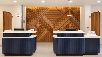 A hotel reception area with two reception desks, a wooden wall with a geometric pattern, and a ceiling with recessed lighting.