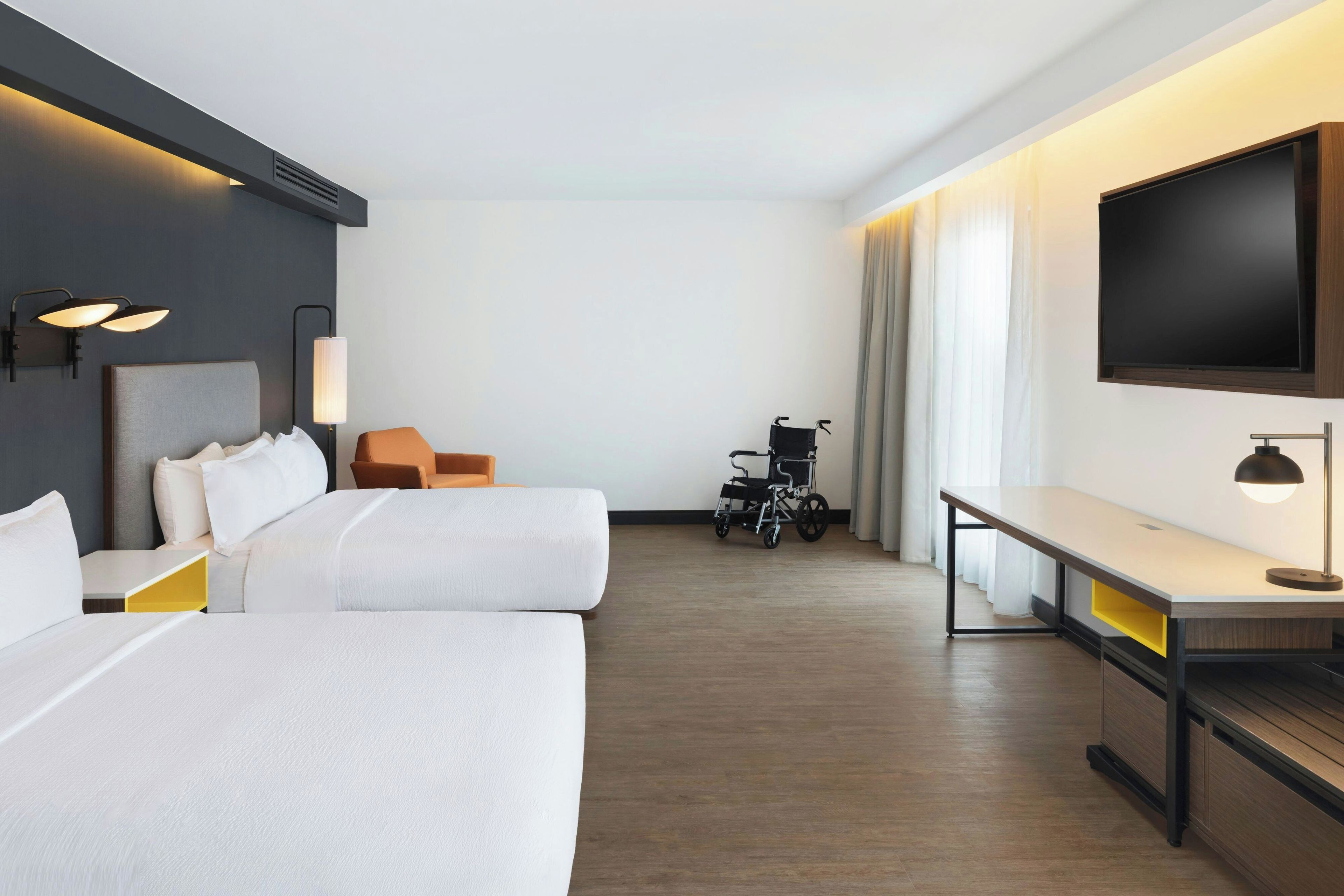 Habitación de hotel con dos camas, un escritorio, una silla, un televisor y un cochecito de bebé.