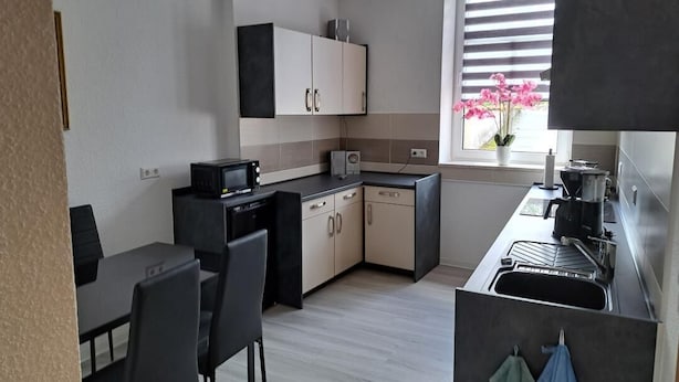 A compact kitchen with a dining area, a window with blinds, and a view of pink flowers outside. - NEW! Apartment Calbe an der Saale (Calbe)