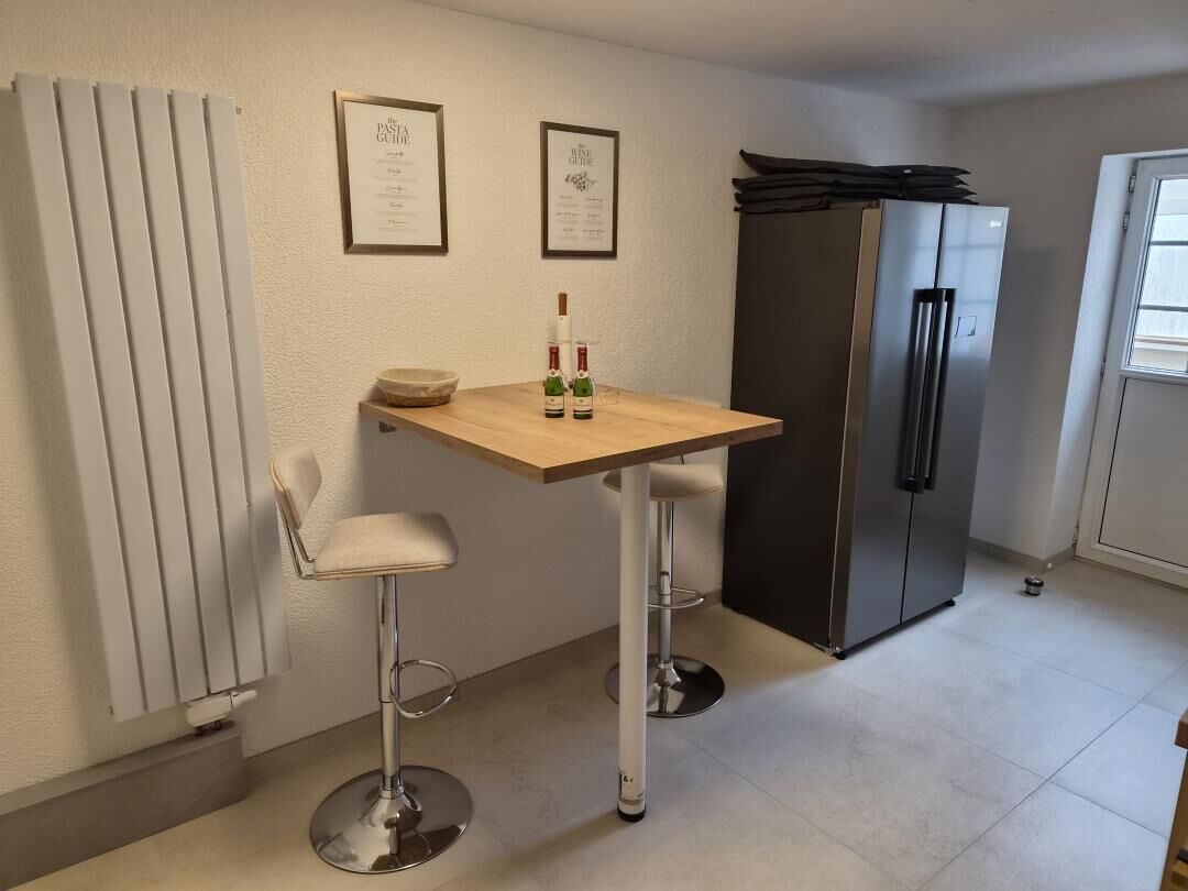 Une cuisine avec une table en bois, deux tabourets de bar, un réfrigérateur et des certificats encadrés accrochés au mur.