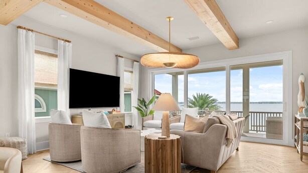 Ein modernes Wohnzimmer mit großem Fernseher, bequemen Sitzmöglichkeiten, einer Holzbalkendecke und Meerblick.
