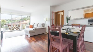 A modern living room with a sofa and chairs, a dining table with chairs, a kitchen area, and a large window offering a view of the outdoors. - Apartamento Calçada Encantada by Atlantic Holiday (Caniço)