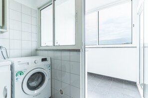 A compact laundry room with a front-loading washing machine, a window, and a tiled floor. - Apartamento Calçada Encantada by Atlantic Holiday (Caniço)
