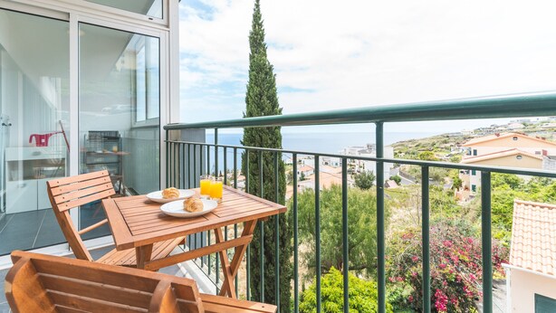 A balcony with a table, chairs, and a view of the sea and buildings. - Apartamento Calçada Encantada by Atlantic Holiday (Caniço)