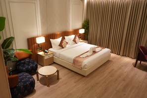 Habitación de hotel con cama, mesitas de noche, lámpara, sillón, una pequeña mesa y una planta en maceta.