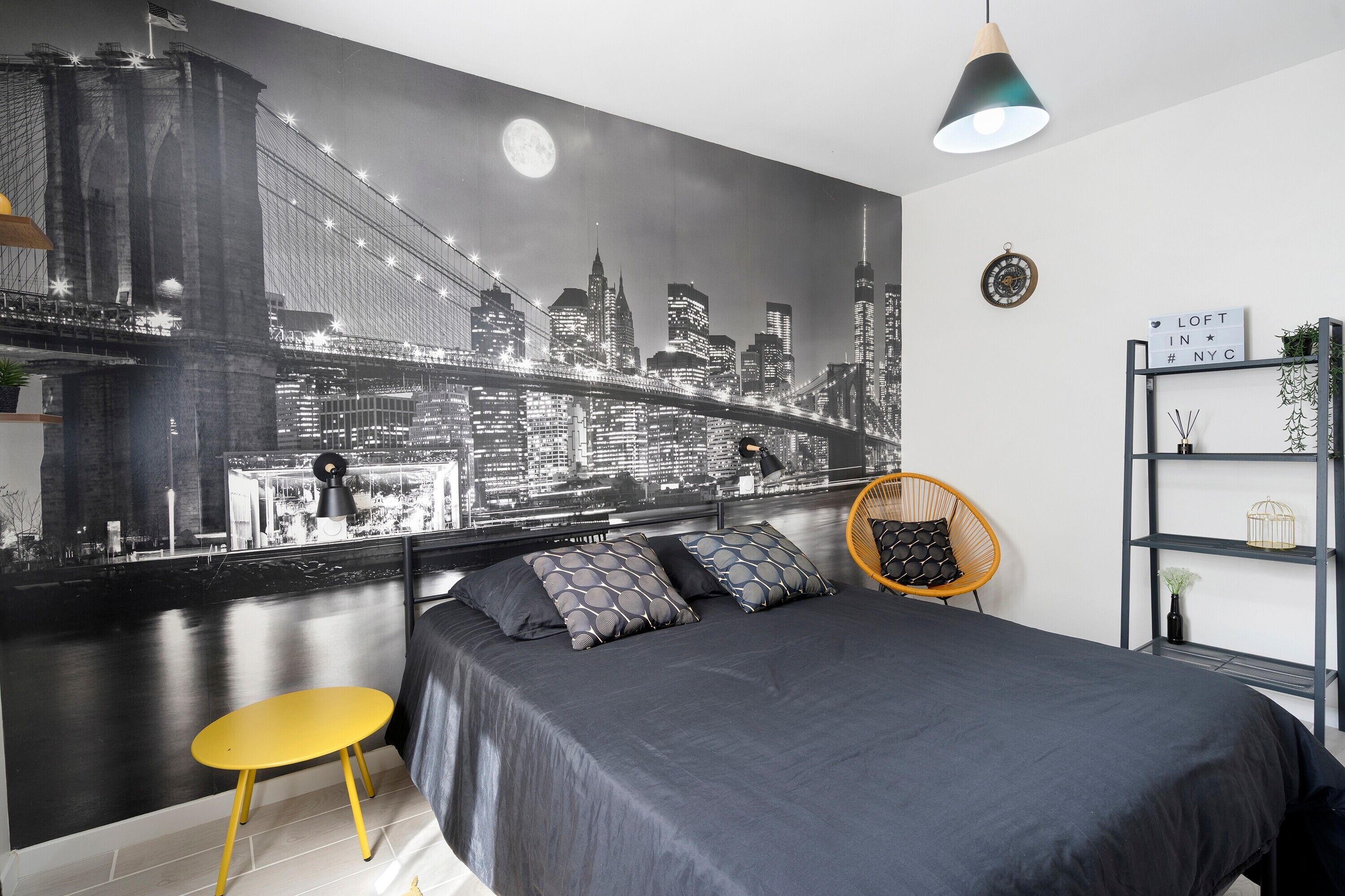 A bedroom with a cityscape mural, a bed, a yellow stool, a hanging chair, and a shelf with decorative items.