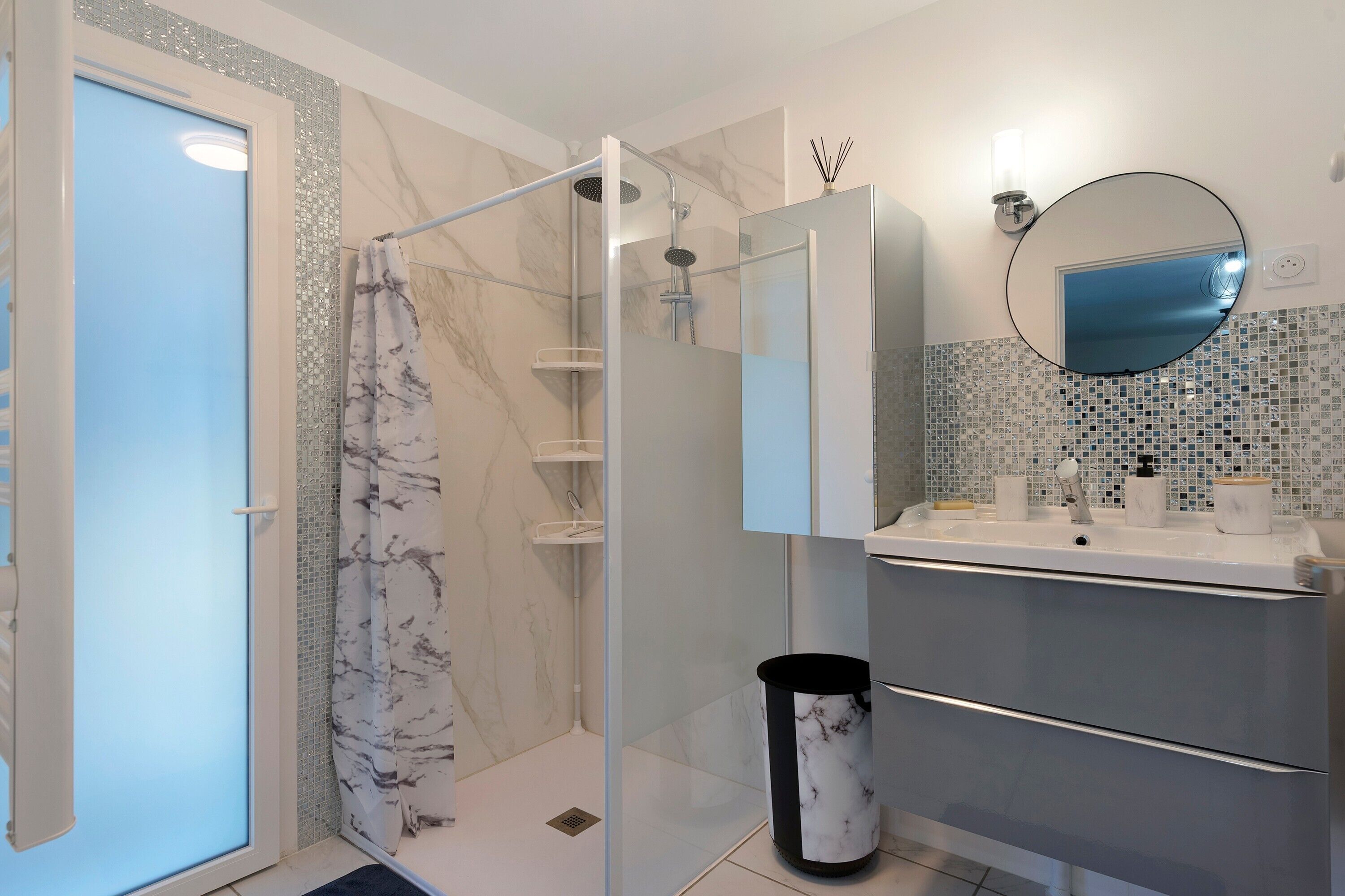 A modern bathroom with a glass shower enclosure, a marble-tiled shower area, a white sink with a mirror, and a tiled backsplash.