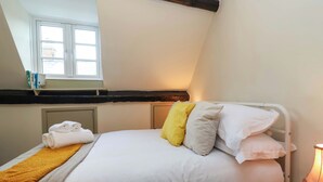 A bedroom with a bed, pillows, a folded towel, a bedside table with a lamp, and a window with books on the sill. - 29 Spring Street (Chipping Norton)