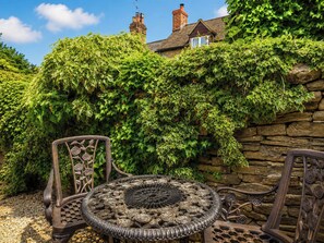 Ein Garten mit einer mit Efeu bewachsenen Steinmauer, einem Metalltisch mit aufwendigen Verzierungen und zwei Metallstühlen mit floralem Dekor.