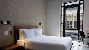 A modern hotel room with a large bed, a nightstand, a wall-mounted light, and a view of the city through a large window.