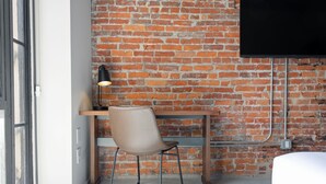 A room with a brick wall, a wooden desk, a chair, a bed, and a TV mounted on the wall.