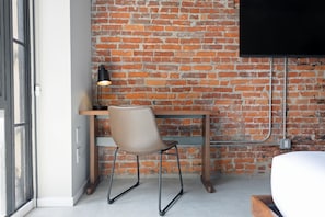 Una habitación con pared de ladrillo, un escritorio de madera, una silla, una cama y un televisor montado en la pared.