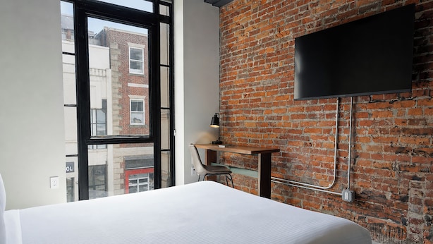 A hotel room with a brick wall, a TV mounted on it, a desk with a chair, and a large window.