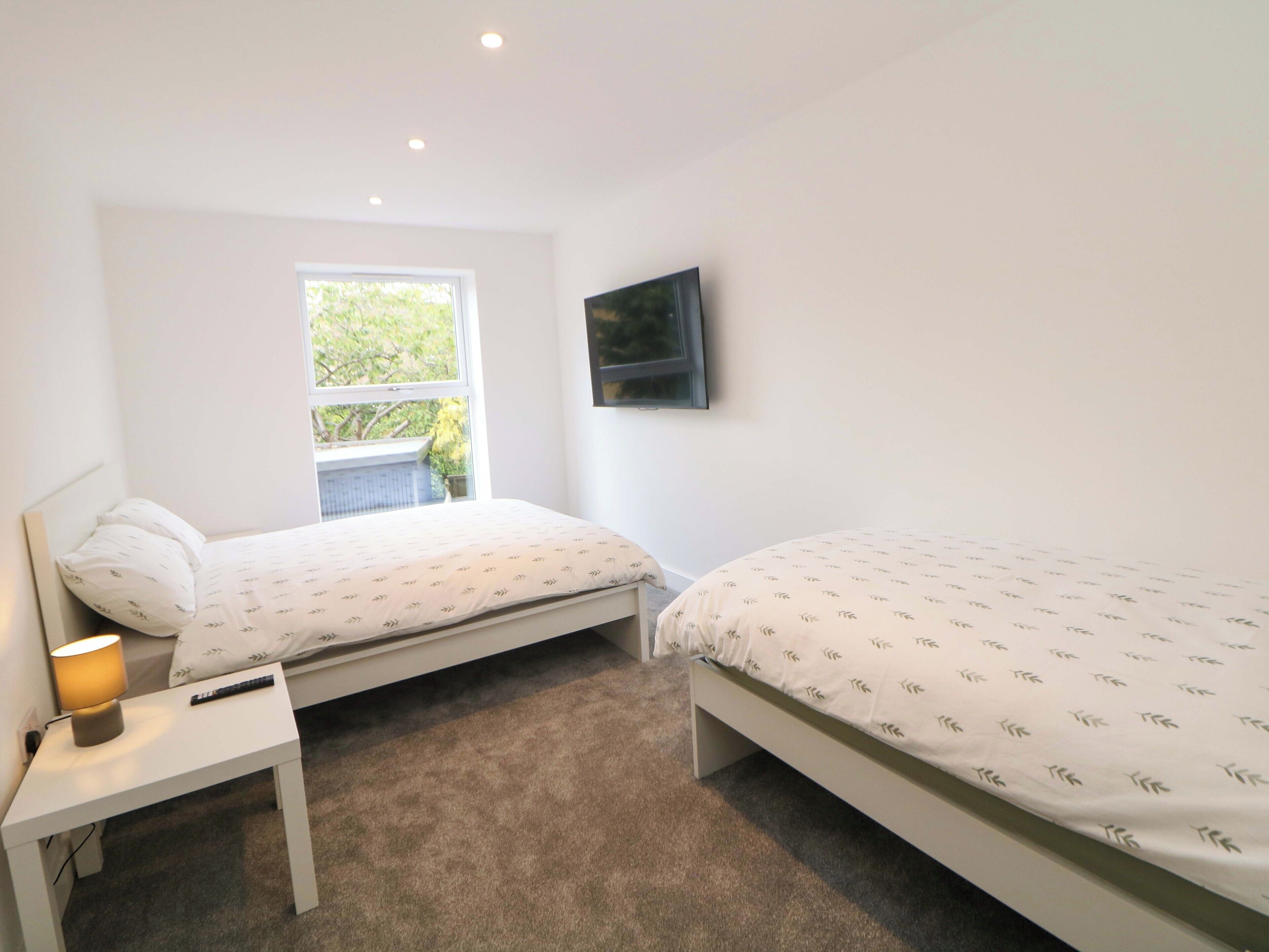 A bedroom with two beds, a TV mounted on the wall, a window, and a bedside table with a lamp.