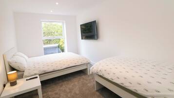 A bedroom with two beds, a TV mounted on the wall, a window, and a bedside table with a lamp.